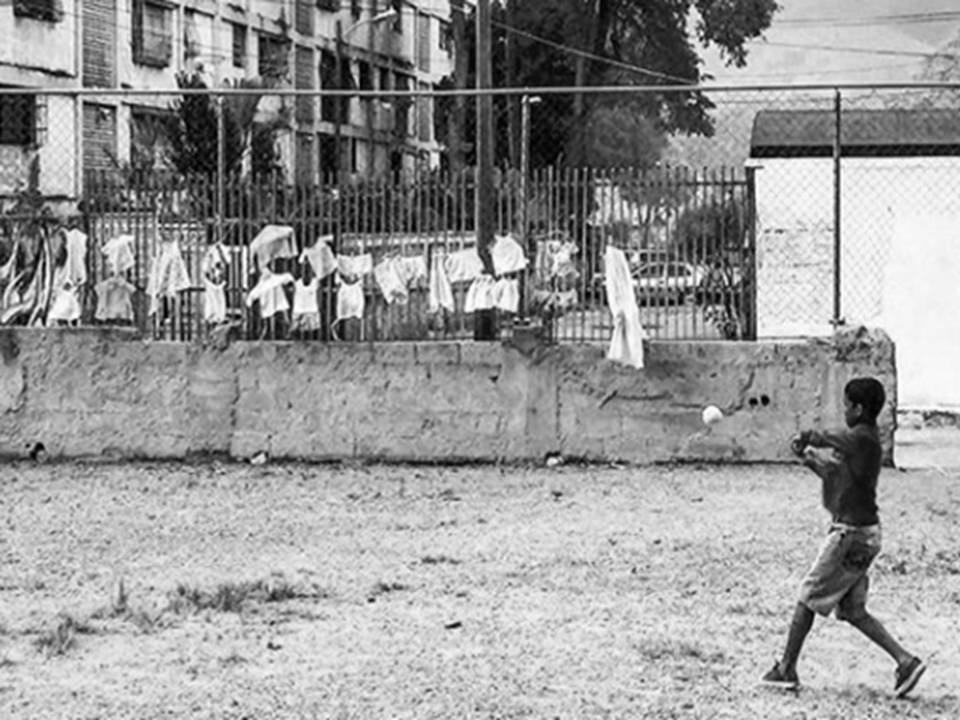 foto blanco y negro de un niño no mayor de 10 años en un terreno baldío, vestido con franela y pantalones cortos sujetando un bate de beisbol en la mano mirando una pelota en el aire. Alrededor bloques de viviendas, un estacionamiento y ropa colgando de las ventanas