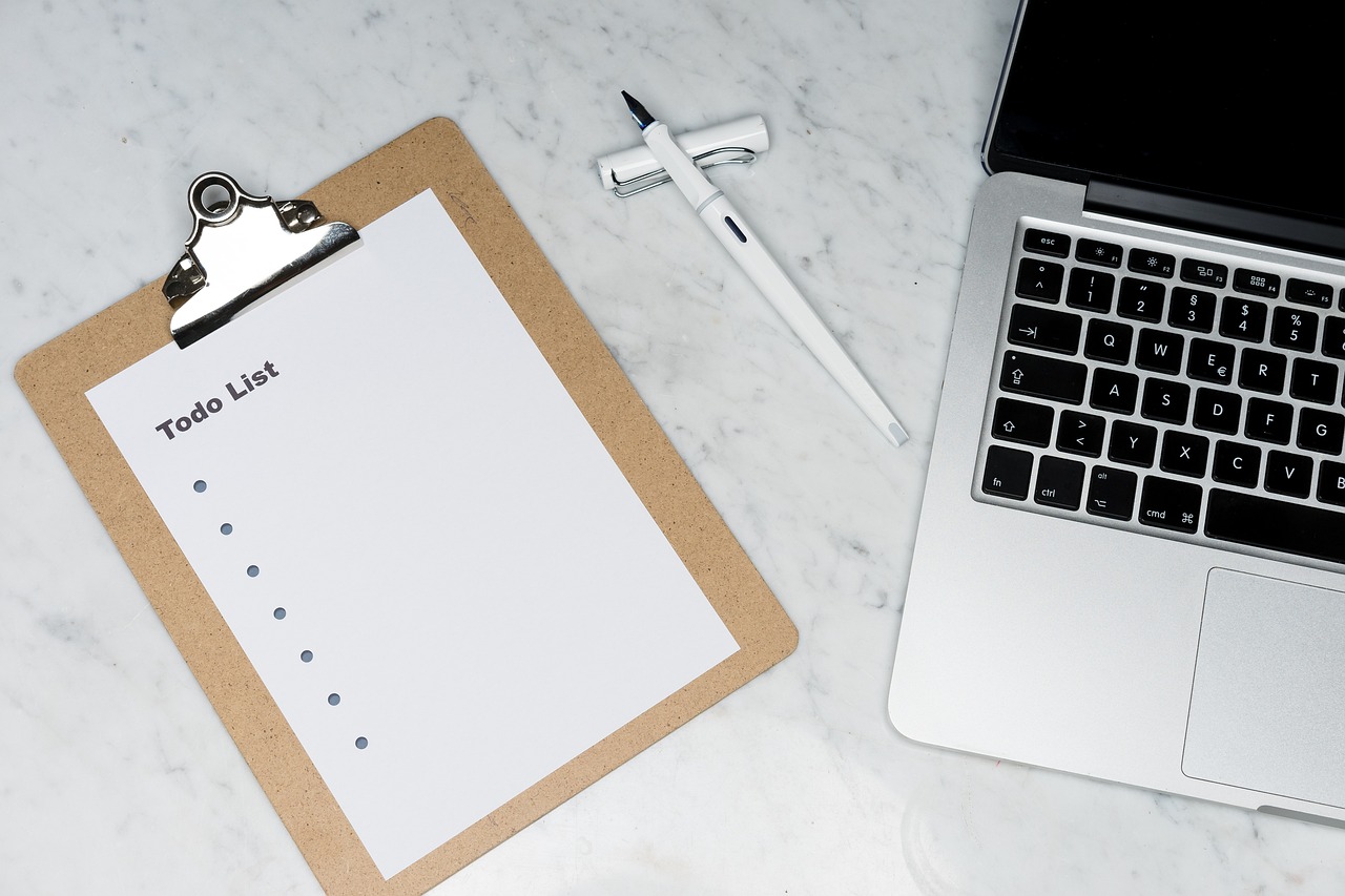 Top-down view of a clipboard labeled “To do List” beside a white pen and a laptop on a marble desk. 
