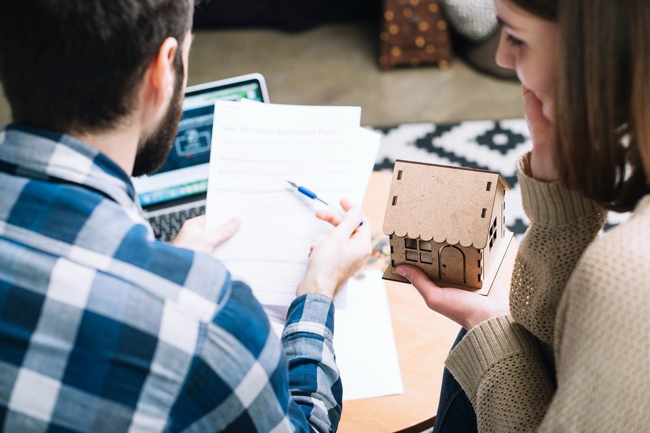 Couple reviews paperwork at a table with a laptop while one person holds a small wooden house model.