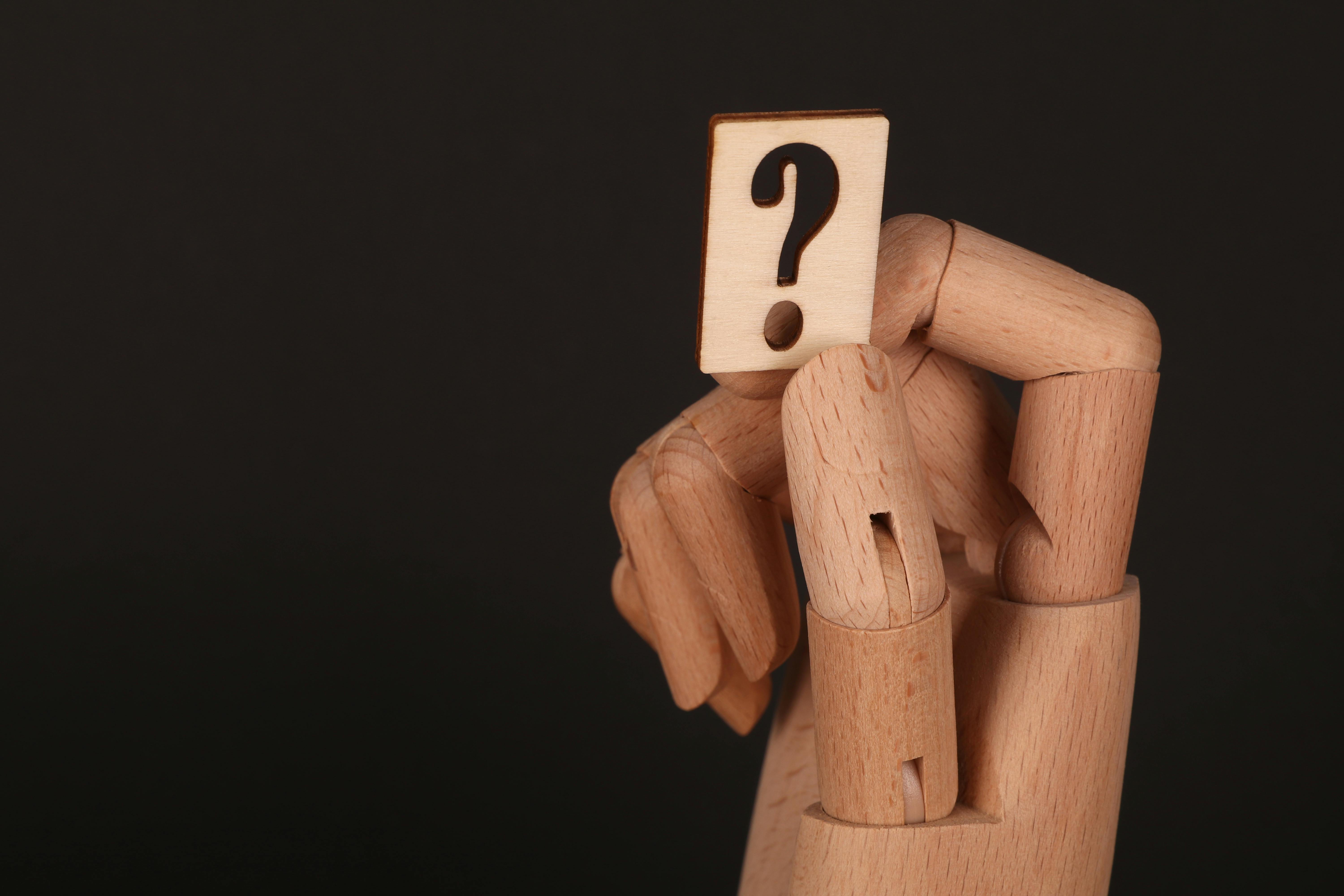 Wooden mannequin-like figure holds its head while a block with a question mark covers its face against a dark background.