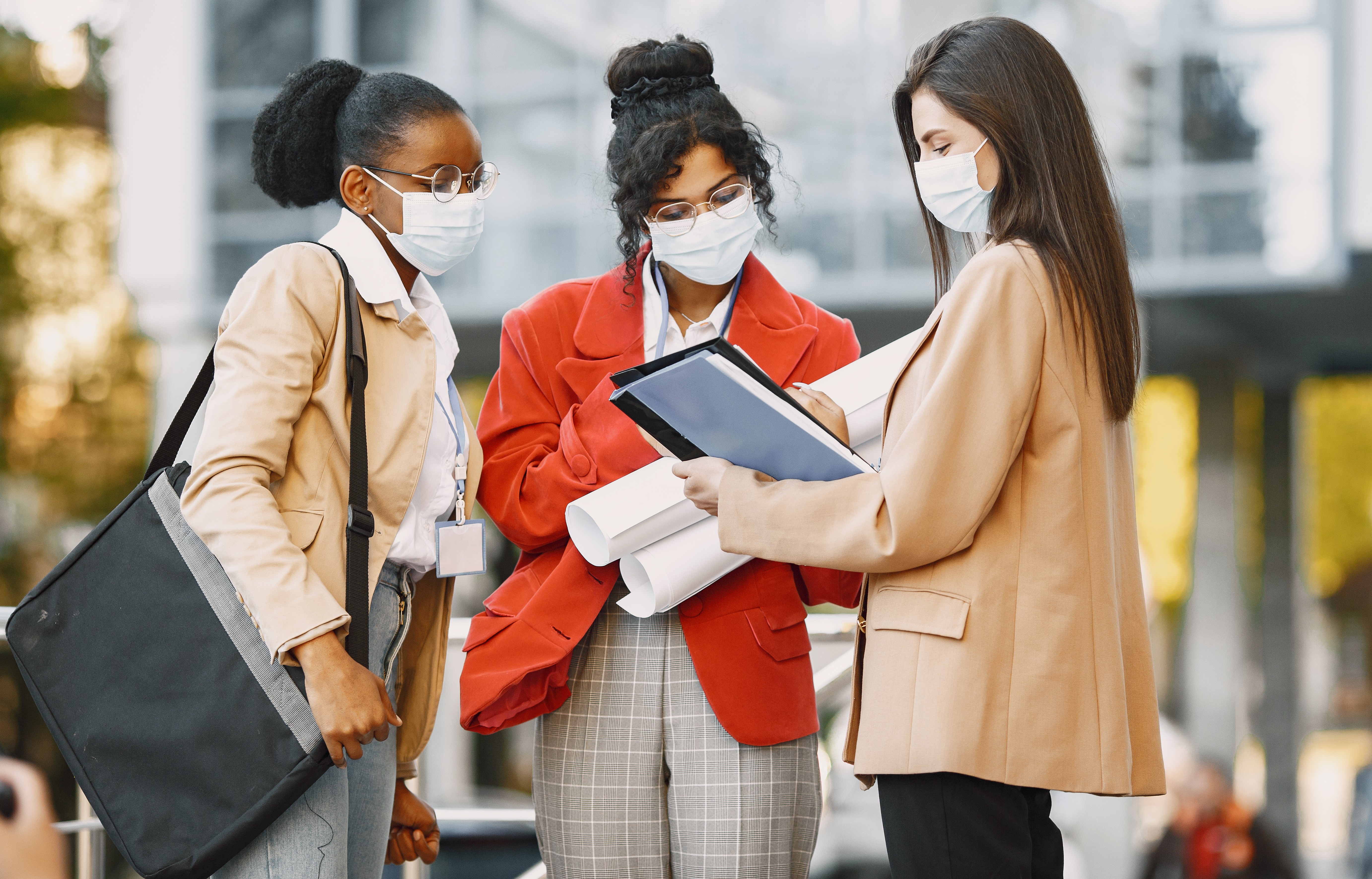 Three women wearing face masks stand outdoors near a modern building, reviewing architectural plans and documents together.