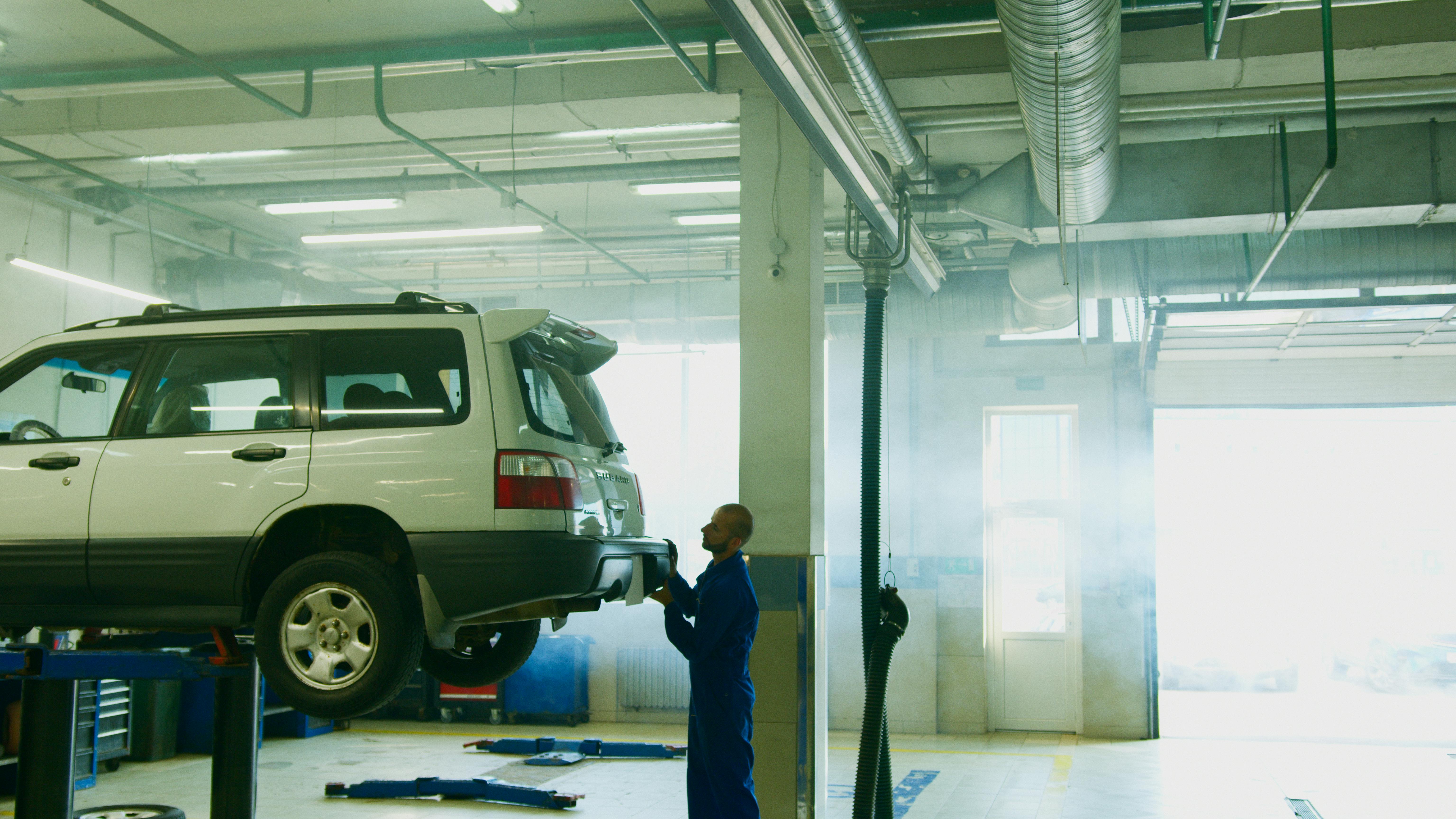 Mechanic in blue coveralls inspects the rear of a white SUV raised on a lift inside a bright, industrial auto repair garage