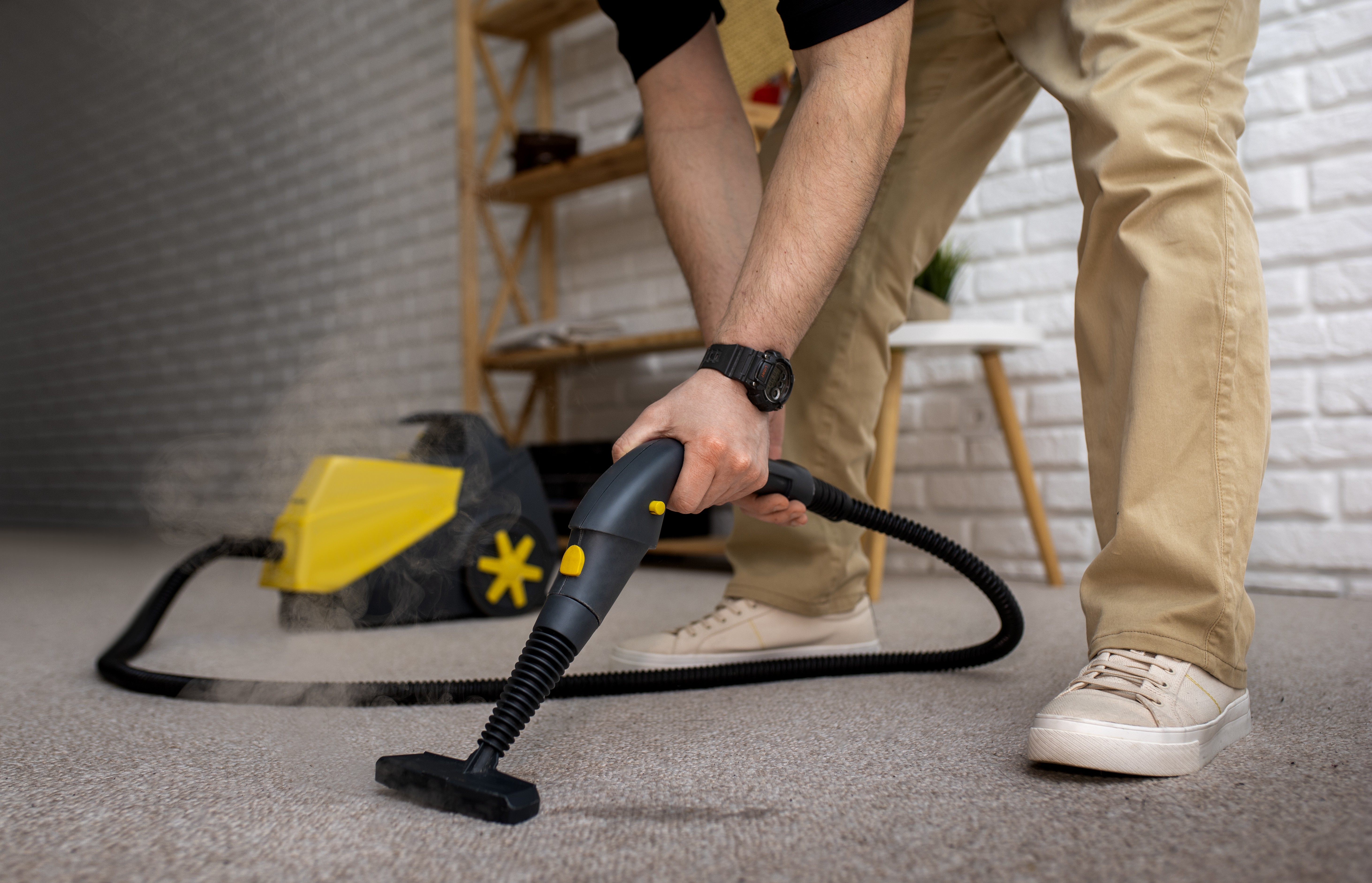 Person using a steam cleaner to deep clean and sanitize a carpet in a living room, with visible steam lifting dirt from the carpet fibers.