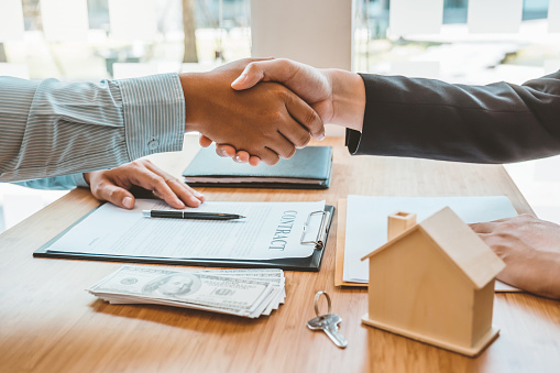 Two people shake hands over a property contract, with cash, keys, and a small model house on the table.