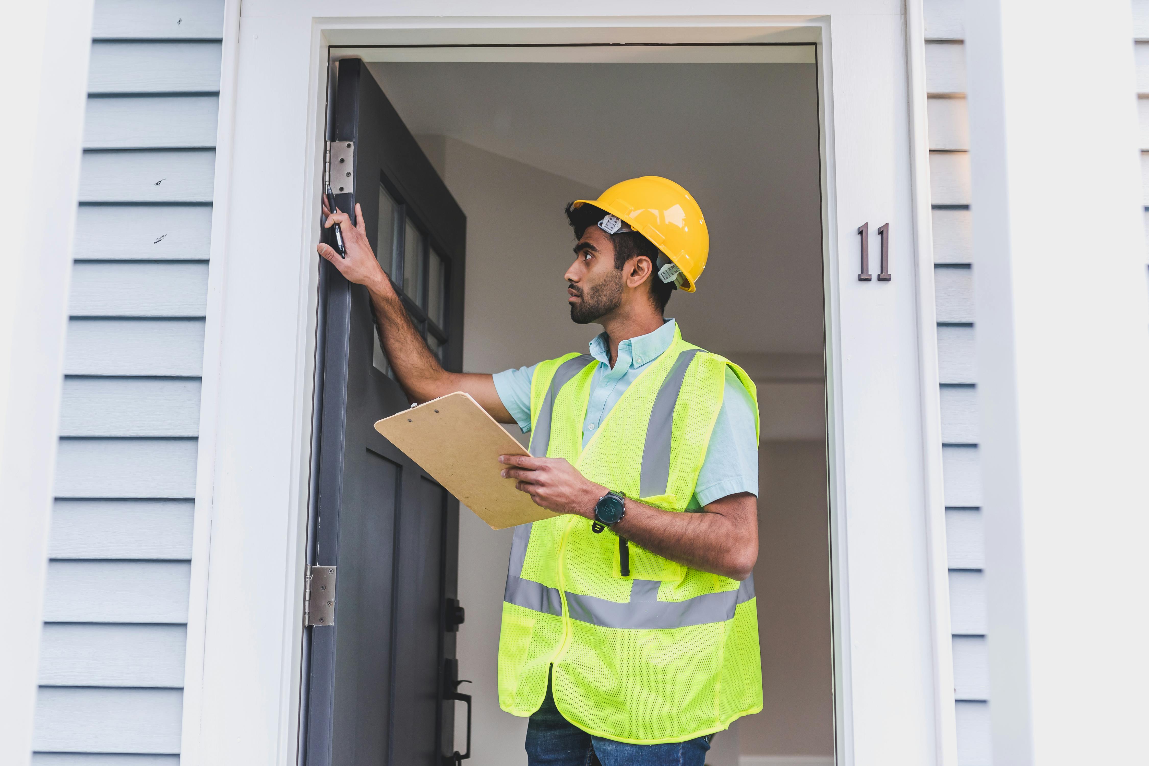 Construction inspector wearing a yellow hard hat and safety vest checking a doorway while holding a clipboard.
