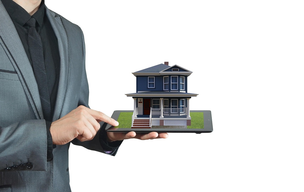 Real estate agent in a gray suit holds a smartphone displaying a small house model on the screen against a white background.