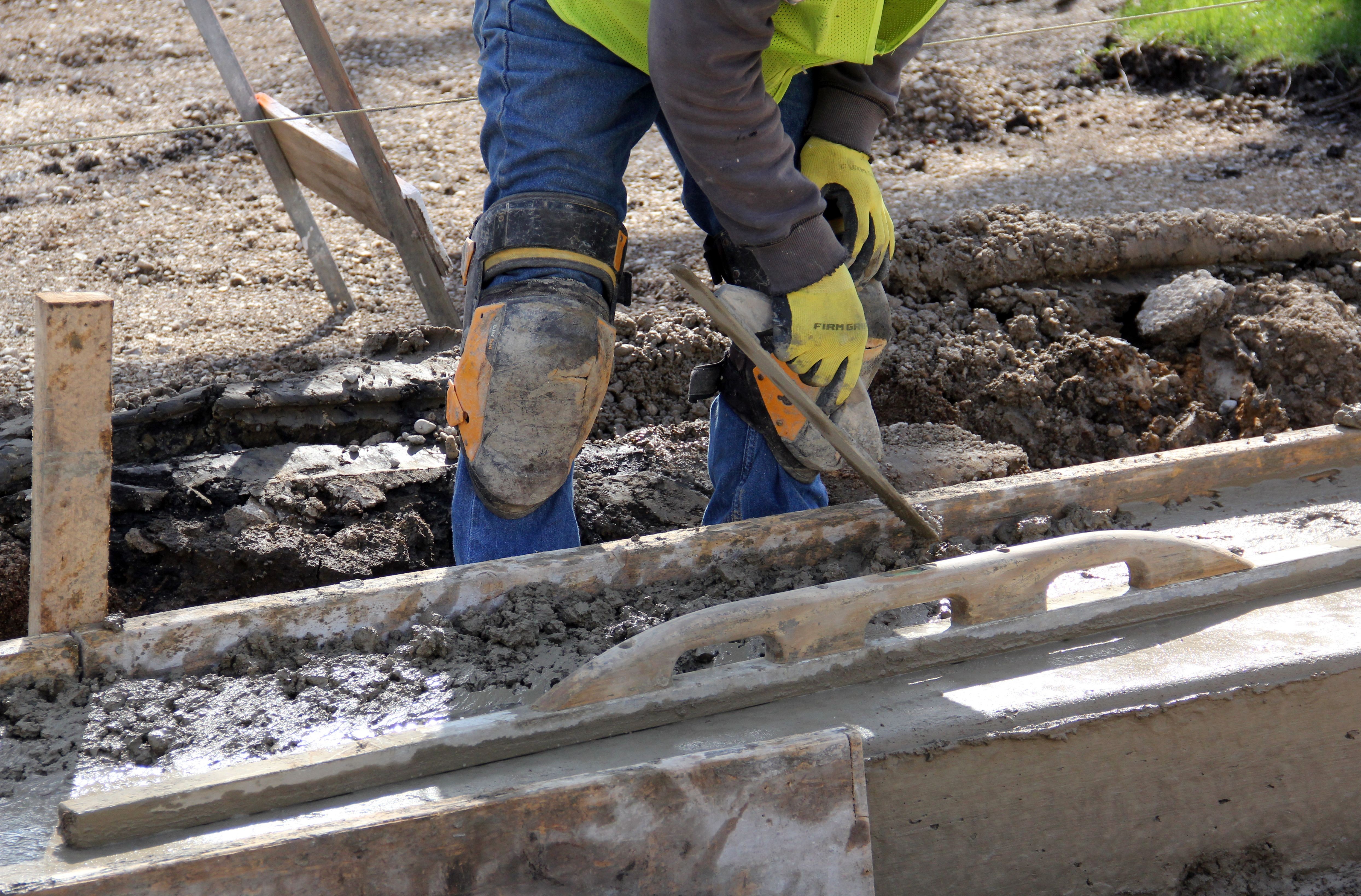 Concrete worker wearing safety gear smoothing wet concrete inside wooden forms at a construction site.