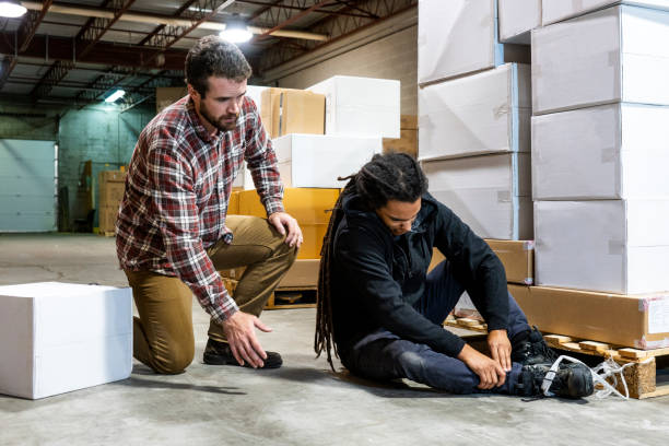A worker kneels beside an injured coworker seated on the warehouse floor near stacked boxes and pallets.