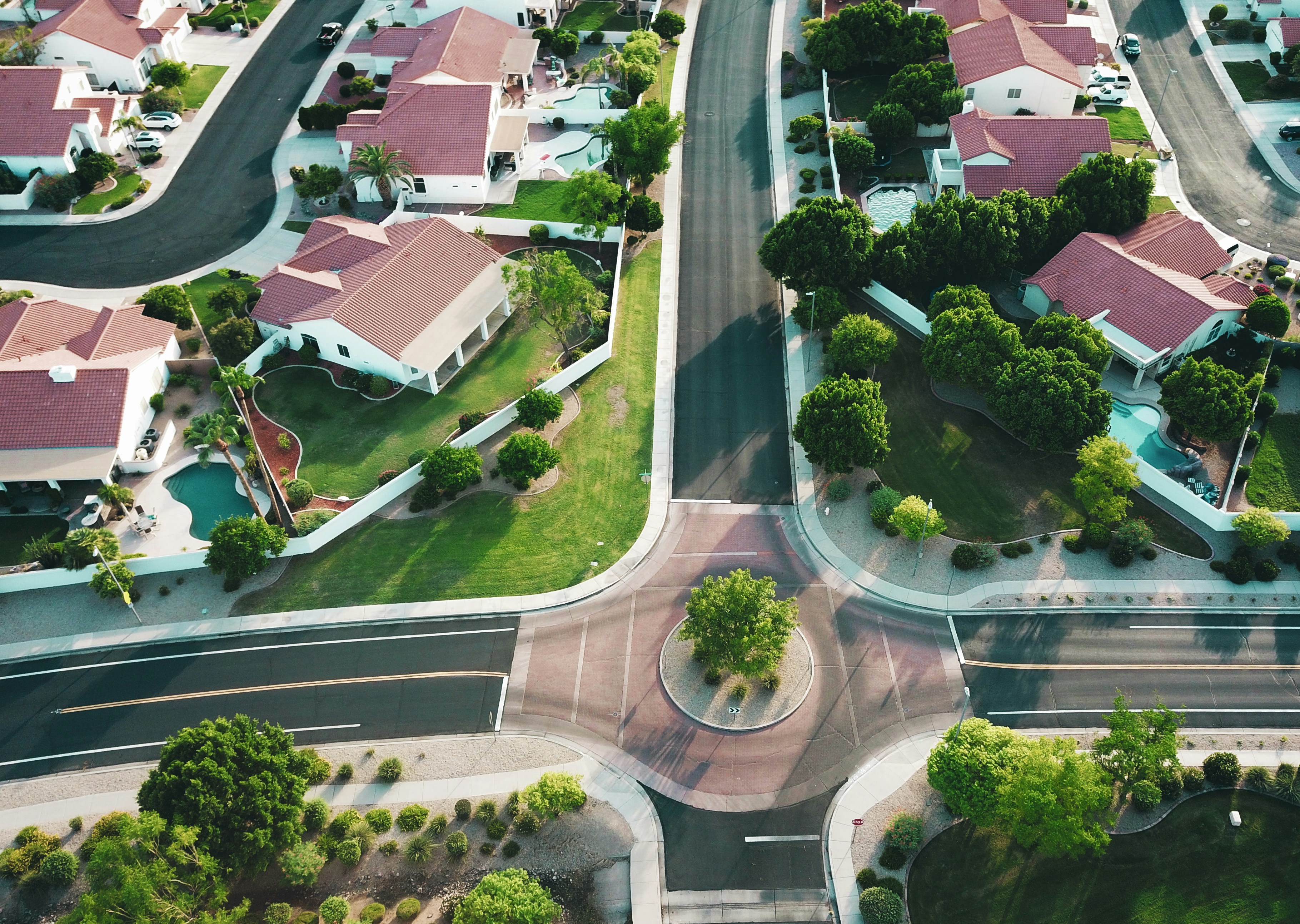 “Aerial view of a suburban neighborhood with red-roofed homes, green yards, and a four-way intersection with a small roundabout.”