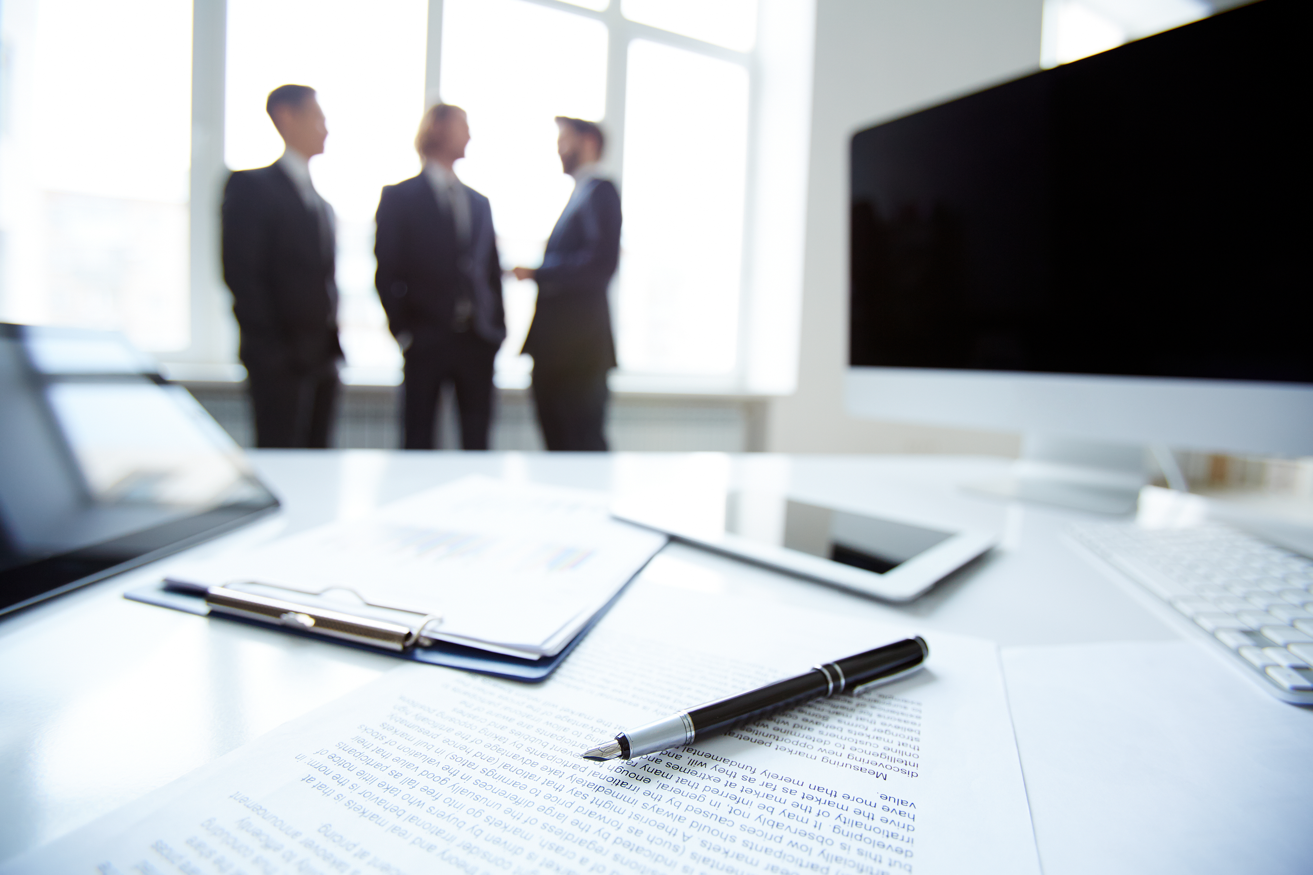 Business documents and a pen on an office desk with professionals meeting in the background.