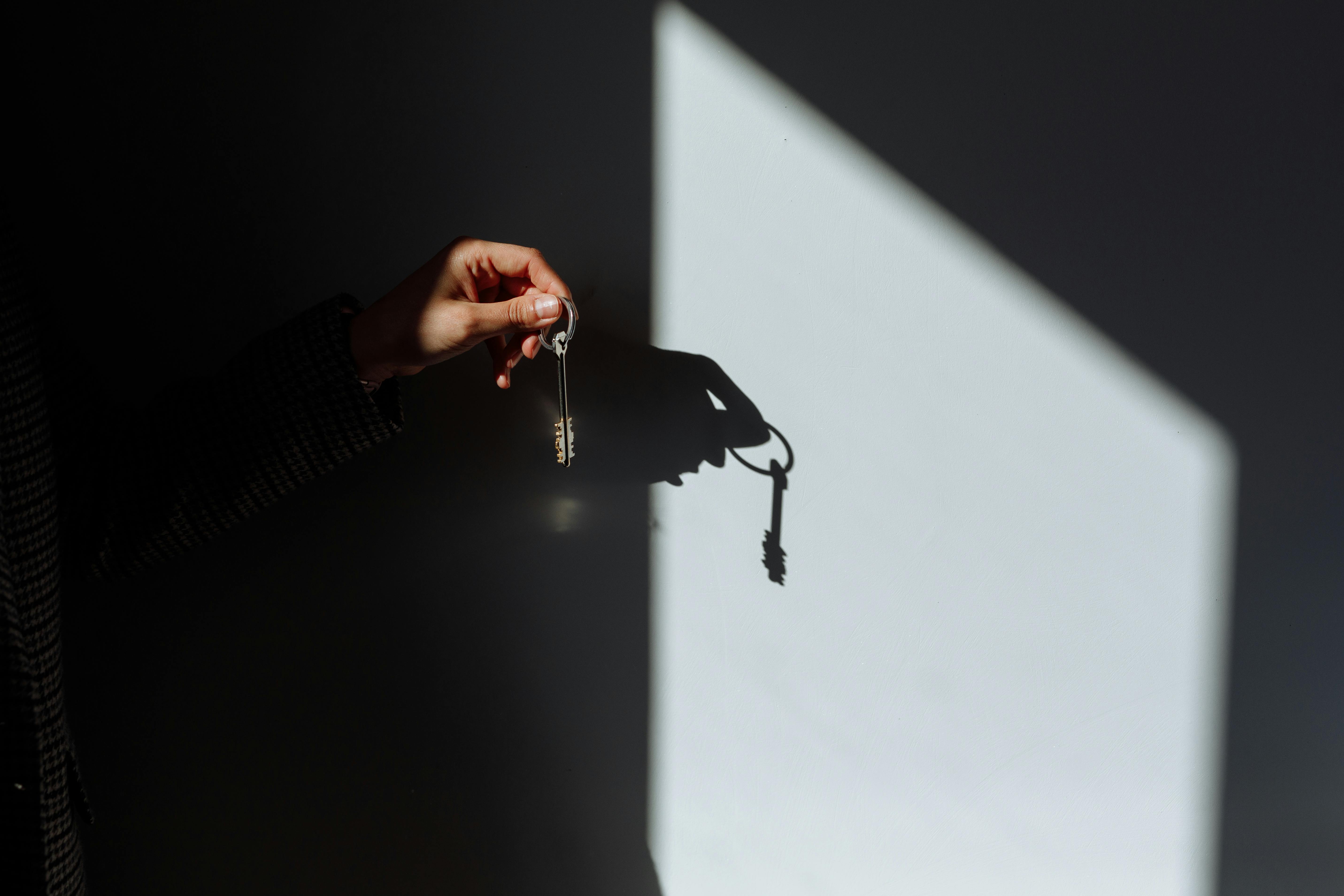 Hand holds a key ring with house keys in front of a wall, casting a sharp shadow in a beam of sunlight.