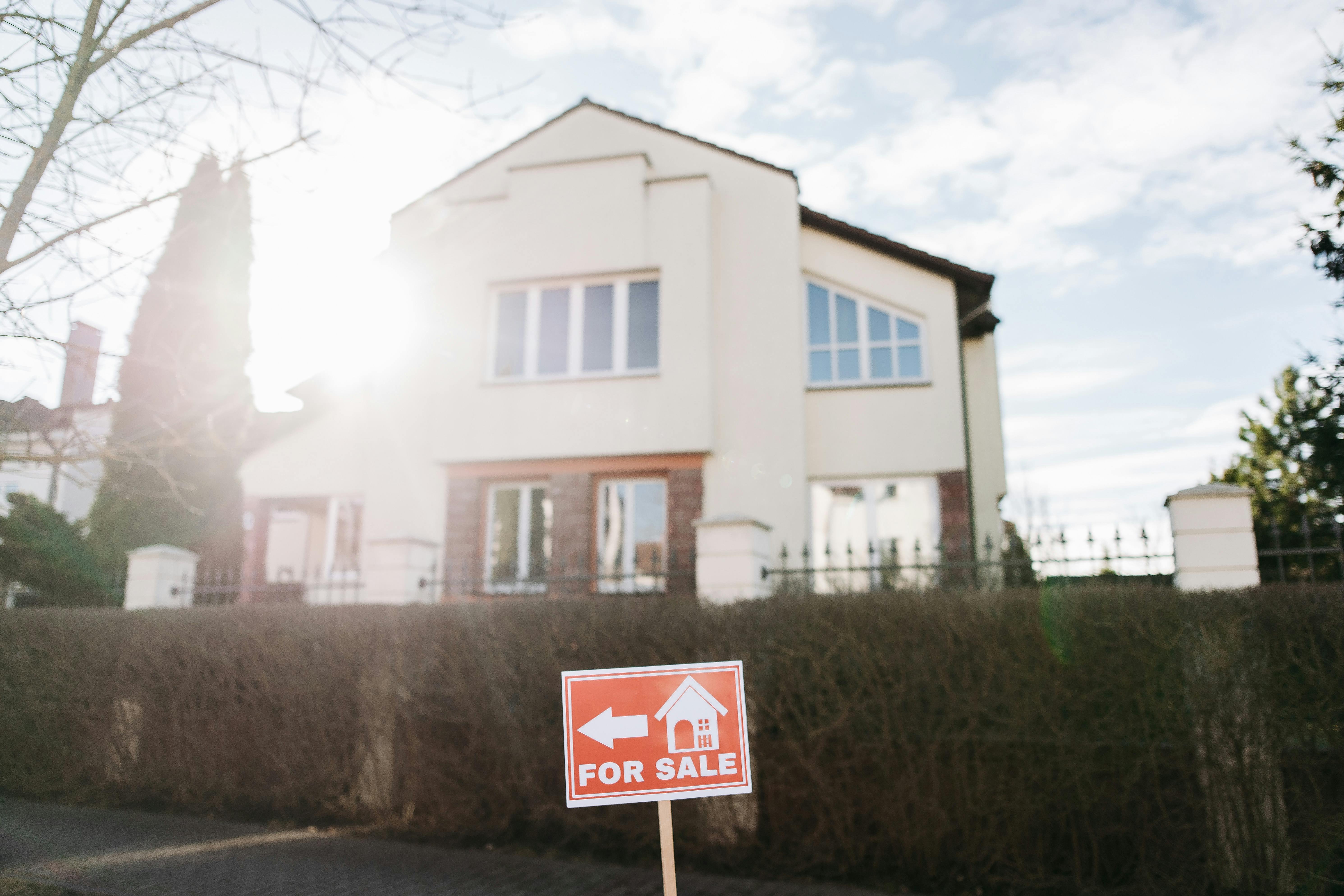 “Red ‘For Sale’ sign in front of a modern two-story house, with bright sunlight flaring across the scene.”
