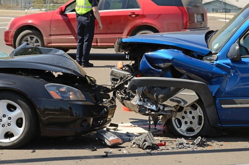 A close-up of a head-on collision between a black car and a blue SUV, both with extensive front-end damage, while an officer manages the accident scene.