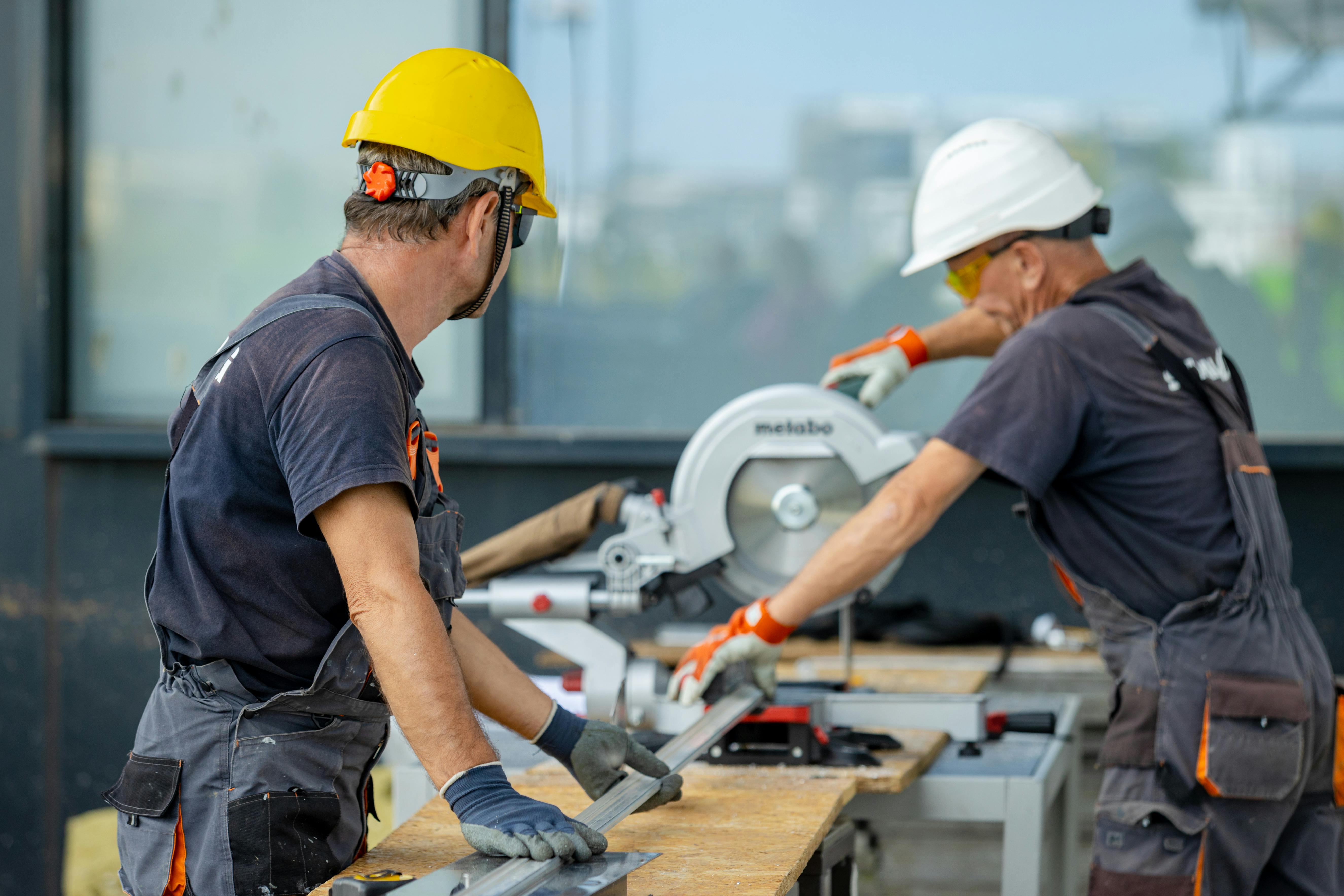 Two construction workers wearing hard hats and safety gear cutting material with a power saw at a job site.