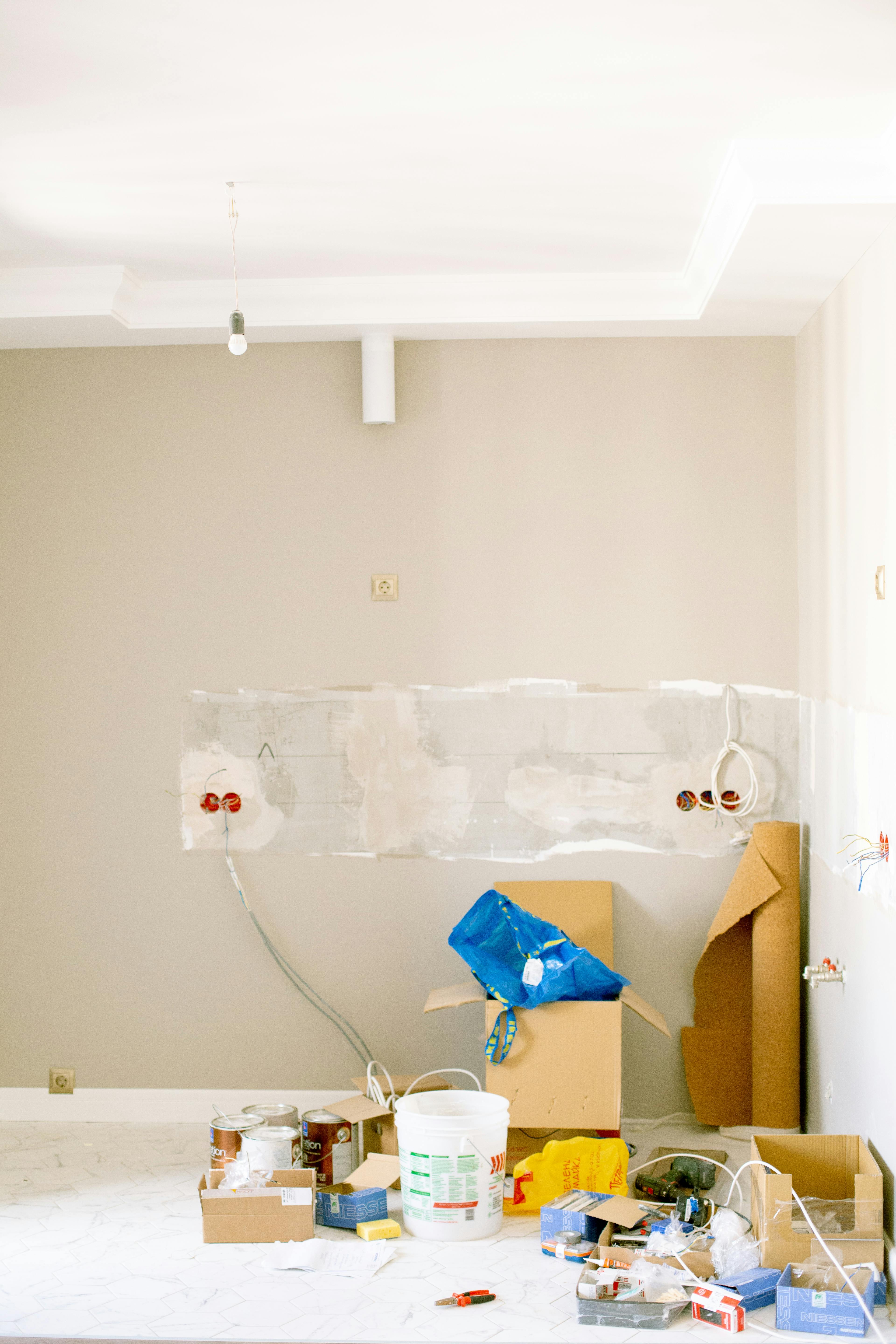 Unfinished room with exposed electrical wiring and renovation supplies scattered on the floor, including paint cans, boxes, and tools.
