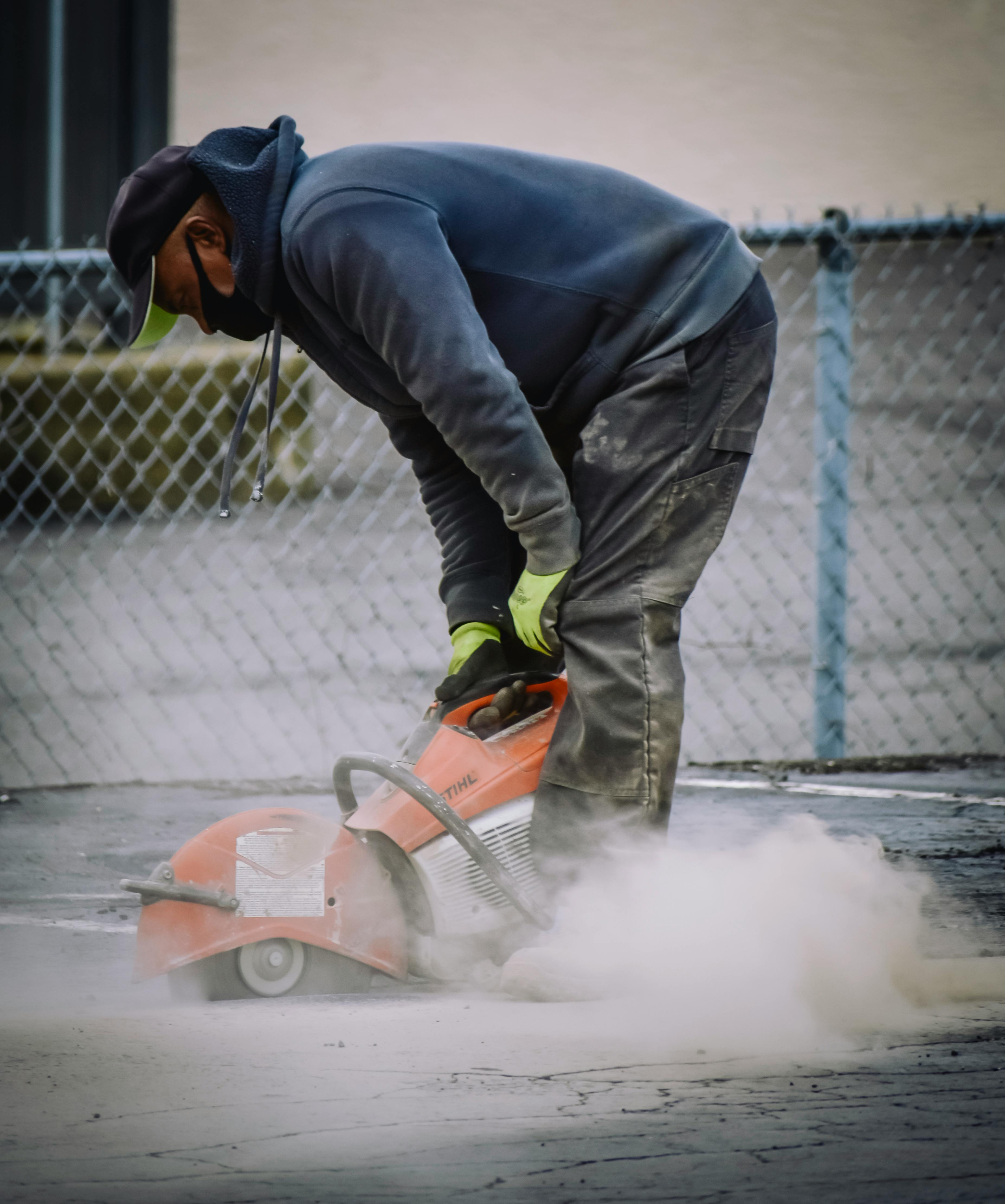 Construction worker using a concrete saw to cut pavement, creating a cloud of dust near a chain-link fence.