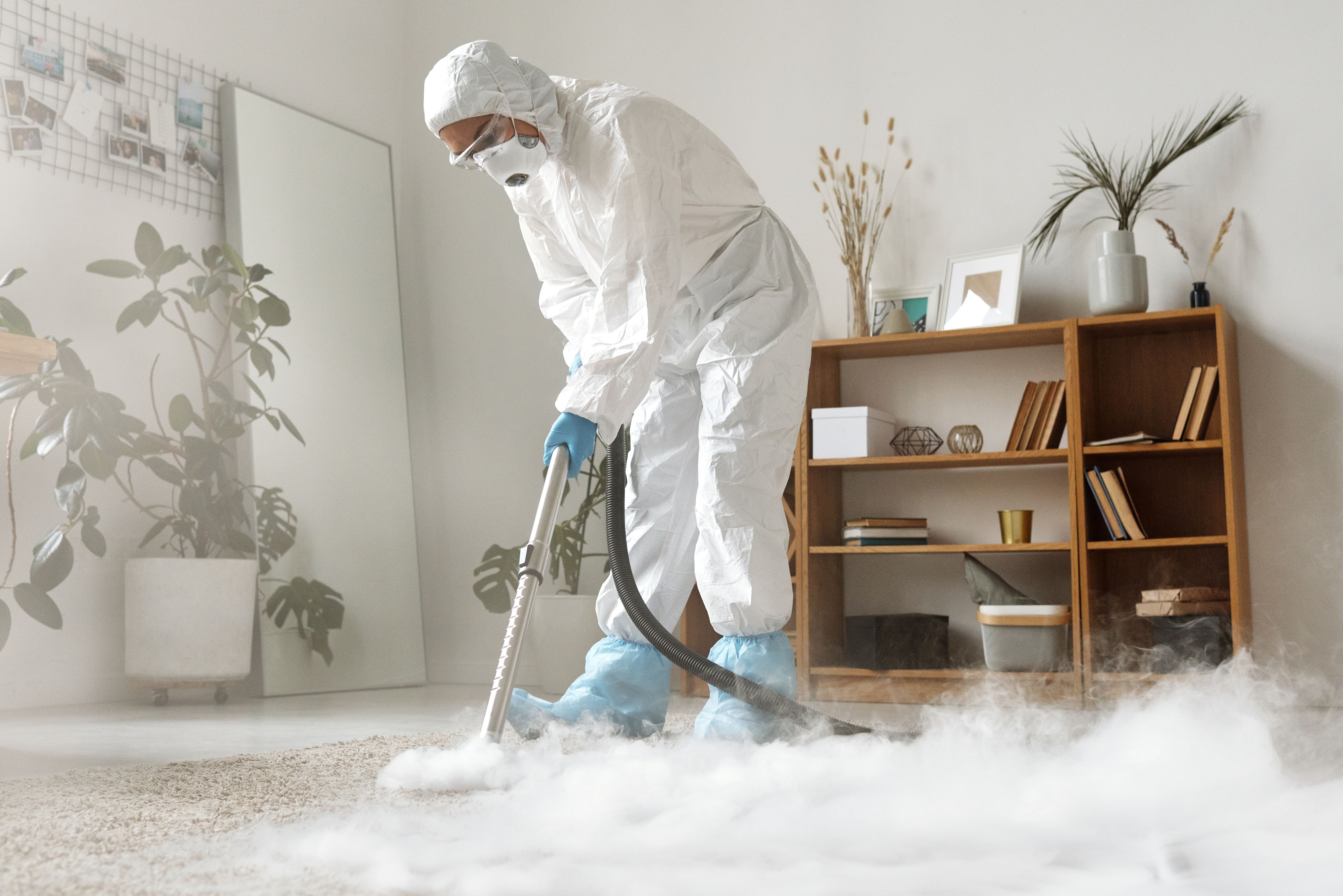 Professional cleaner wearing full protective gear and a respirator uses a vacuum to clean and disinfect a carpet in a residential room, with cleaning mist visible around the work area.