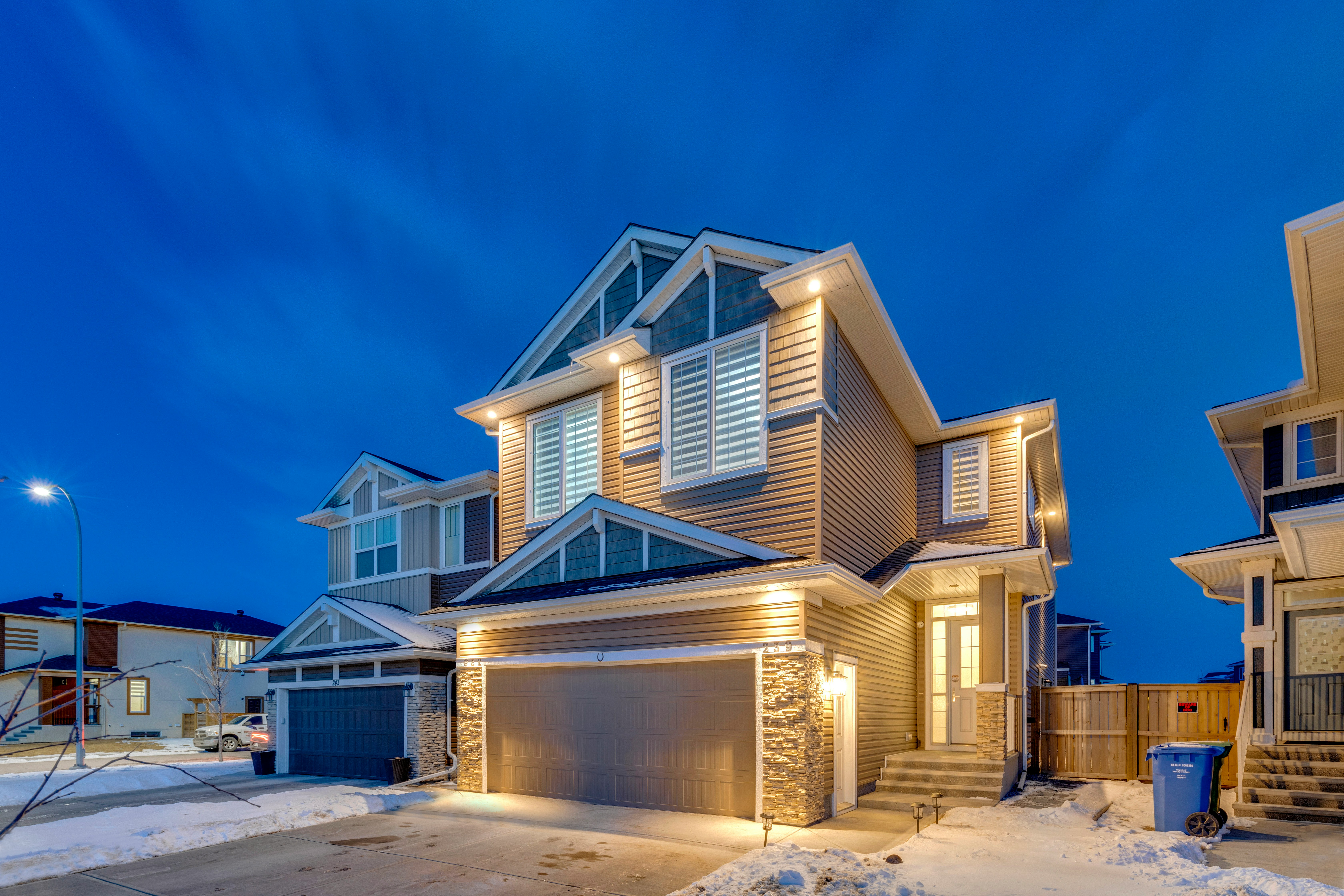 A two-story suburban house with exterior lights on at dusk, surrounded by snow and neighboring homes.