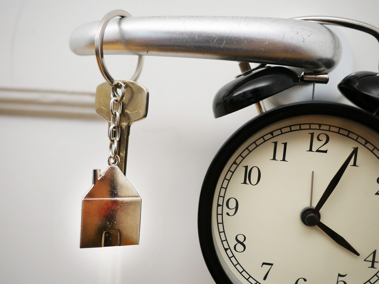 Close-up of a keychain with a house-shaped keyring hanging next to a black alarm clock, symbolizing home deadlines or move-in time.