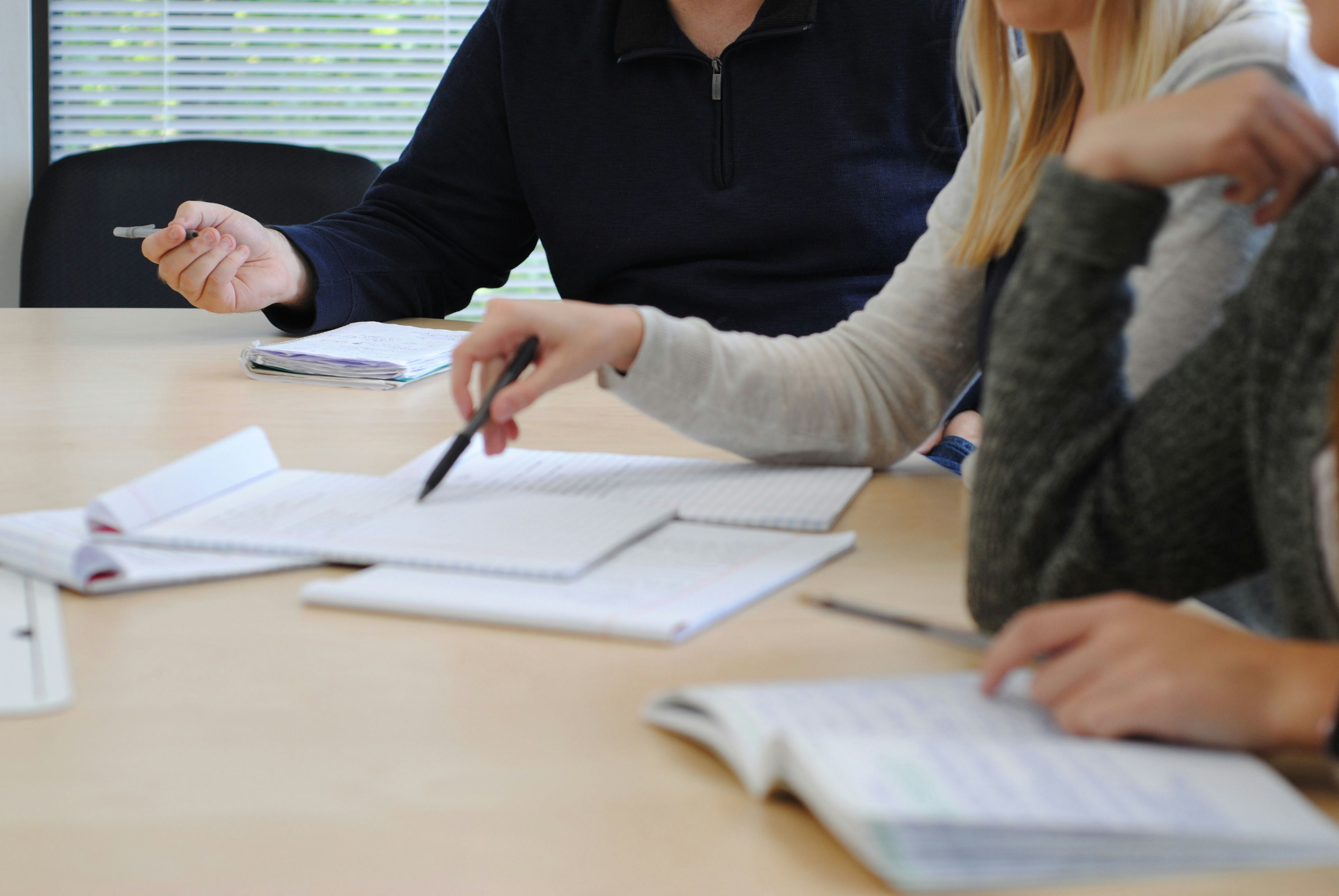Three people sit around a table reviewing notebooks and written materials, with one person pointing to a page during a collaborative discussion or study session.