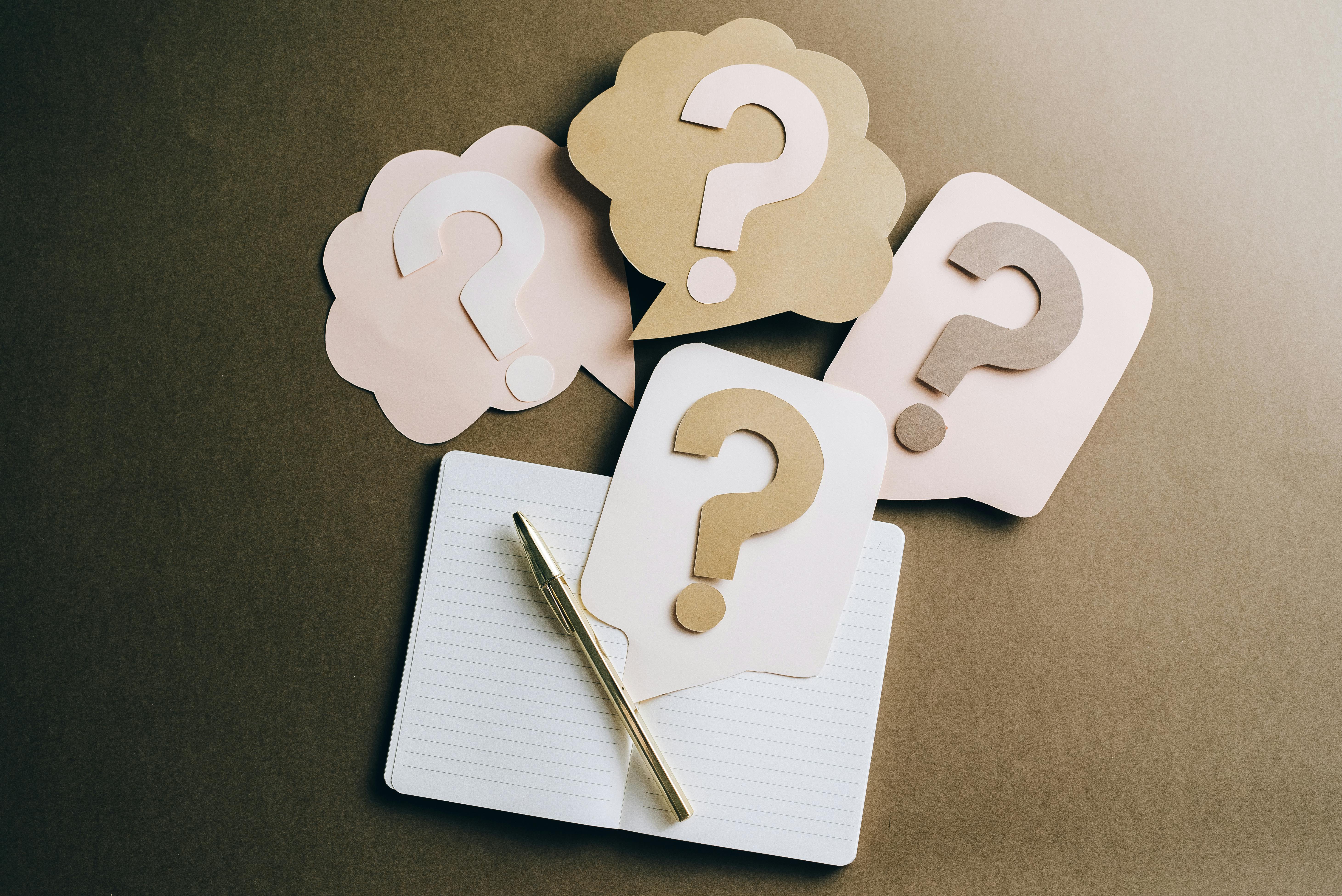 Paper cutout question marks arranged above an open notebook with a pen, representing uncertainty or FAQs.