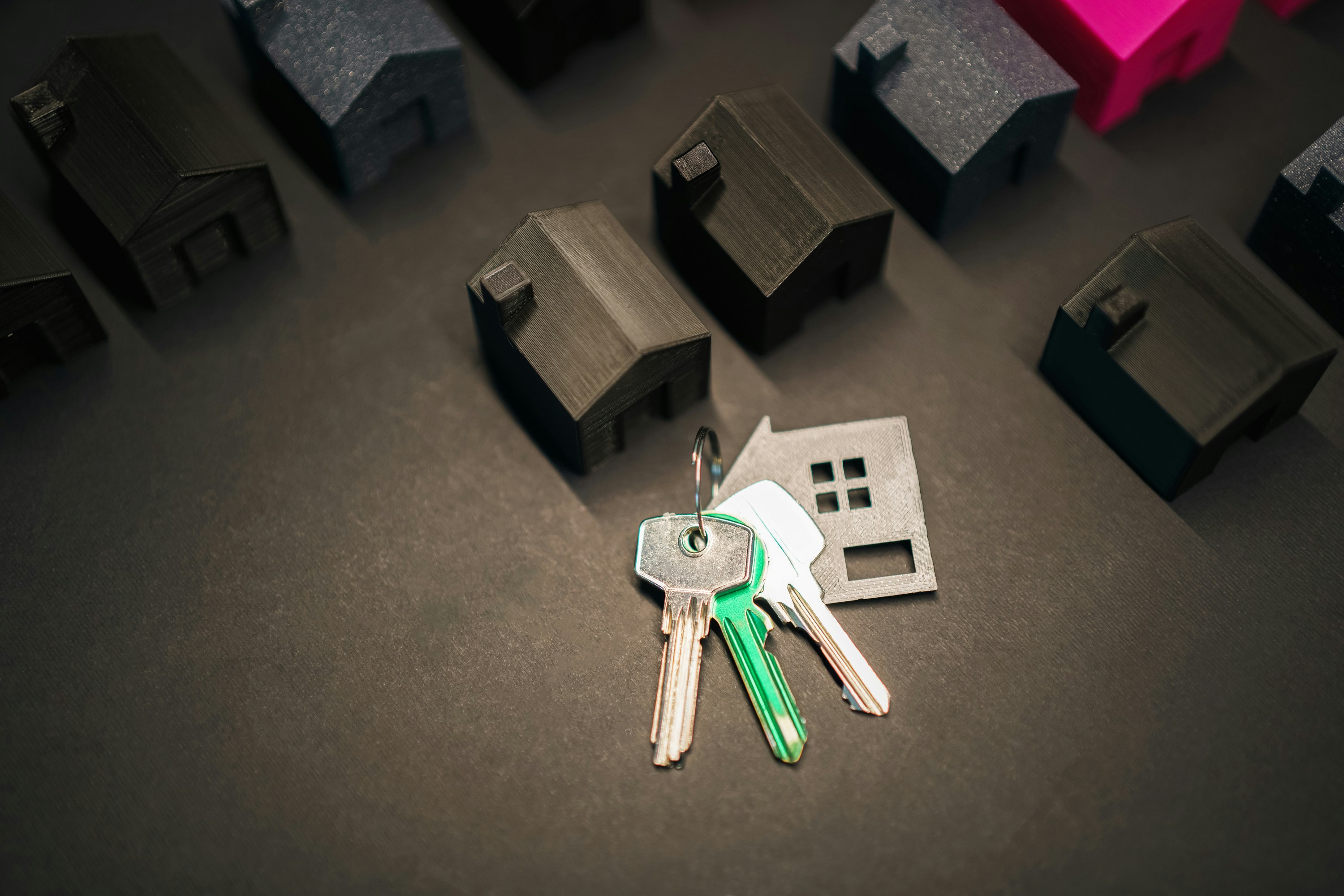 “House-shaped keychain and a set of keys on a dark surface beside miniature model houses, symbolizing buying or selling a home.”